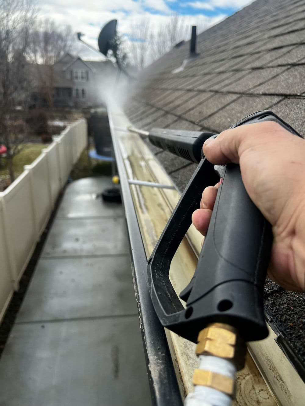 Person using a pressure washer to clean roof gutters with a residential background.